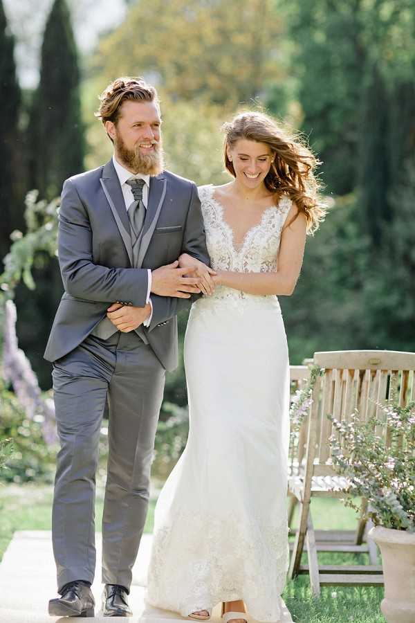 A couple portrait taken outdoors in a garden setting, with the bride and groom walking arm in arm and laughing. The groom wears a charcoal grey three-piece suit with a grey tie and black dress shoes, and has a beard. The bride wears a fitted ivory lace wedding gown with a deep V-neckline, lace cap sleeves, and a mermaid silhouette, with her hair down and wind-blown. A wooden garden bench and a terracotta pot with soft lavender-toned florals are visible in the background. The image is a mid-length portrait shot with soft natural light and a blurred garden backdrop featuring cypress trees.