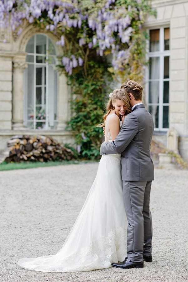 A couple portrait taken outdoors on a gravel courtyard in front of a classic French chateau facade covered in blooming purple wisteria. The bride wears a white lace ball gown with a cathedral-length train and has her hair half-up with a small floral hair accessory, while the groom wears a gray suit with a dark tie and black dress shoes. The groom embraces the bride from behind as she smiles and leans into him. The composition is a full-length portrait shot with the chateau's arched windows and ivy-covered stone walls softly blurred in the background. Potential venue feature image.