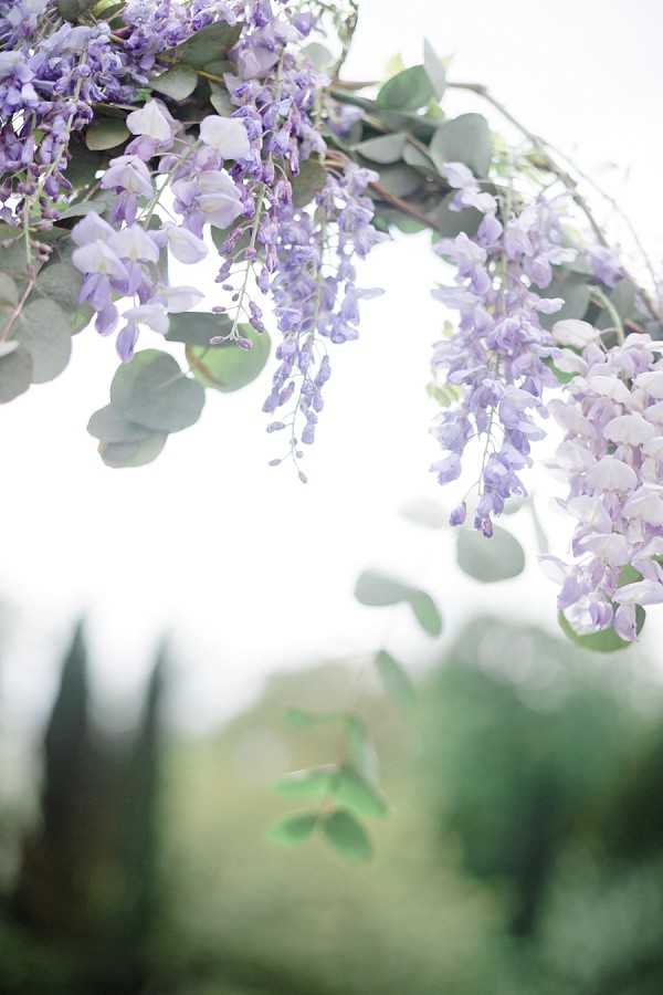 Close-up detail shot of a floral arch or overhead installation featuring cascading lavender and soft purple wisteria clusters mixed with silver-dollar eucalyptus leaves. The background is heavily blurred, revealing an outdoor garden setting with cypress tree silhouettes visible in the soft-focus distance. The composition is shot from below looking up, with bright, overexposed natural light creating an airy, high-key effect. The floral palette is entirely in soft purple and lavender tones paired with muted green eucalyptus foliage, suggesting a romantic, garden-inspired ceremony styling.