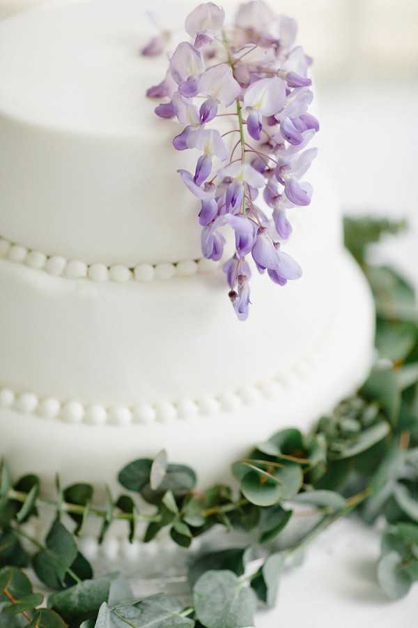 Close-up detail shot of a two-tier white wedding cake with a smooth fondant finish and delicate pearl bead trim along the base of each tier. The upper tier is decorated with a cascading cluster of fresh lilac wisteria blooms. The base of the cake is surrounded by trailing eucalyptus greenery. The overall styling palette is white, soft purple, and green, suggesting a classic yet garden-inspired aesthetic.