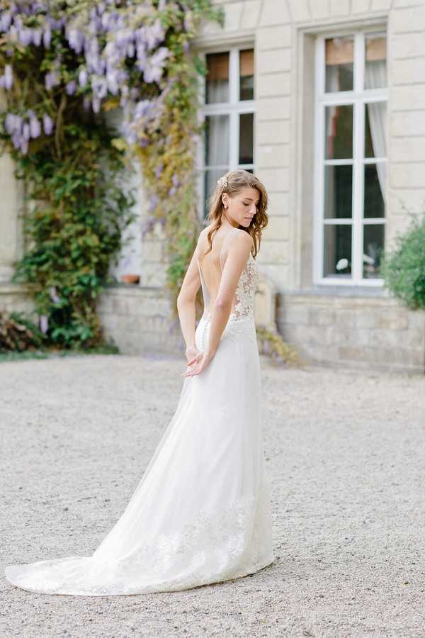 A bridal portrait shot outdoors in the gravel courtyard of a French chateau, with the bride photographed from behind and in three-quarter profile as she glances downward over her shoulder. She wears an ivory A-line wedding gown with a deep open back, lace-appliqué bodice and hem detailing, and a flowing tulle skirt with a short train. Her hair is styled in a soft updo with a small floral or jeweled hair accessory. The chateau facade with tall white-framed windows is visible in the background, partially draped in wisteria with lavender blooms. The overall styling is classic and refined, with a light, airy feel. Medium portrait composition shot in natural daylight. Potential venue feature image.