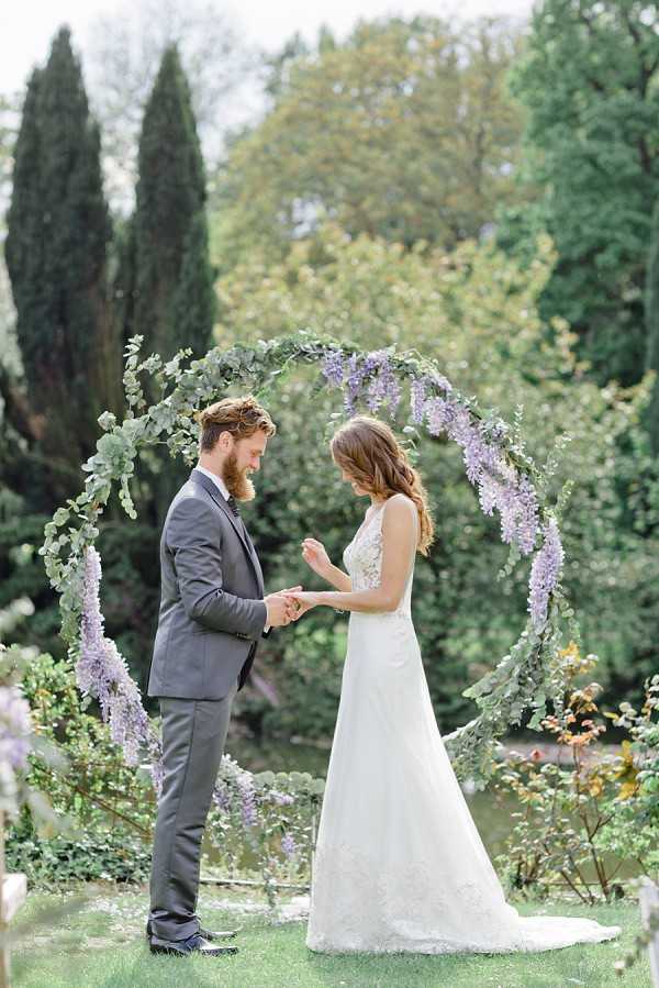 A couple exchanges rings during an outdoor garden ceremony, standing beneath a circular arch decorated with trailing lavender wisteria and eucalyptus greenery. The groom wears a charcoal grey suit and places a ring on the bride's finger while both look down at their hands. The bride wears a sleeveless A-line lace gown with a short train. The setting is a lush garden with cypress trees and dense foliage in the background, giving the scene a romantic garden aesthetic. The image is a medium full-length portrait shot with soft, airy natural lighting.