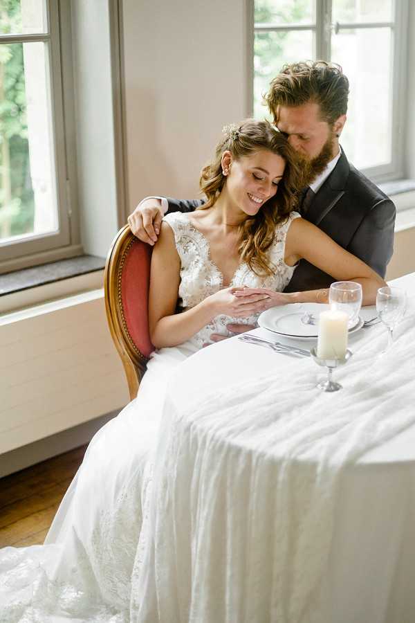 A couple poses at a sweetheart table in an intimate indoor reception or styled shoot setting, photographed in a medium portrait shot. The bride is seated in a vintage-style wooden chair with a red upholstered back, wearing a white lace gown with a deep V-neckline and floral appliqué detailing, her wavy brown hair loosely styled with a small floral hair accessory. The groom stands behind her in a dark charcoal suit, leaning in close with his arm around her shoulder. The table is dressed with a white linen cloth and set with a white plate, silver cutlery, a clear wine glass, and a white pillar candle in a glass holder. Natural window light fills the room from large French-style windows in the background, and the floor is warm hardwood. The overall styling is classic with romantic touches.