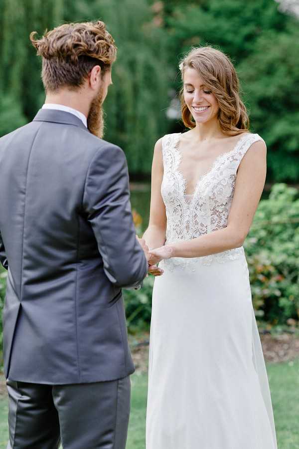 A ring exchange moment between a bride and groom during an outdoor ceremony, set against a lush green garden backdrop. The bride is wearing a white gown with a deep V-neck lace bodice and a flowy chiffon skirt, with her hair loosely styled and a small white floral hair accessory; she is smiling with her eyes downcast as the groom places a ring on her finger. The groom is seen from behind, wearing a charcoal grey suit with a light blue dress shirt. The shot is a medium close-up portrait focused on the couple, with the garden softly blurred in the background.