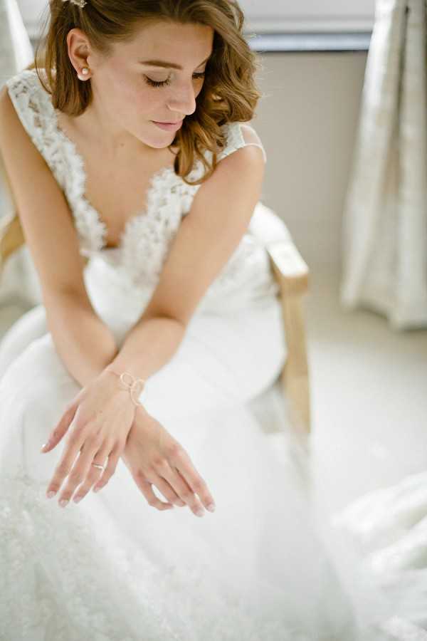 Close-up portrait of a bride seated in a wooden chair in an indoor getting-ready setting, looking down with eyes cast downward in a quiet, reflective moment. She wears an ivory lace wedding gown with a deep V-neckline and lace cap sleeves, paired with pearl stud earrings, a delicate gold hand-chain bracelet, and a thin gold ring. Her dark auburn hair is styled in soft, loose waves with a small hair accessory at the crown, and her makeup is natural with defined lashes. The shot is taken from a slightly elevated angle, emphasizing her hands and the full skirt of the dress, with soft natural light coming from a window in the background.