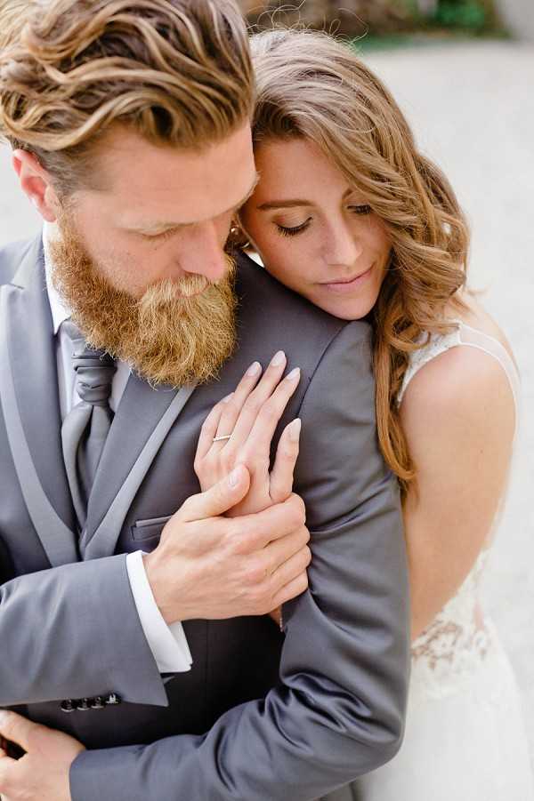 A close-up couple portrait showing the bride embracing the groom from behind, her hands clasped over his chest with a wedding ring visible on her finger. The groom wears a charcoal grey suit with a matching grey tie and white dress shirt, and has a full auburn beard and wavy highlighted hair. The bride wears a lace-detailed ivory dress with an open back, and has loose wavy brunette hair with natural makeup. Both have their eyes cast downward in a quiet, intimate pose. The shot is tightly framed from roughly the shoulders up, drawing focus to the hands and the couple's closeness.