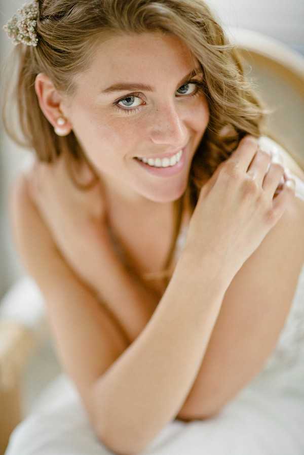 Close-up portrait of a bride during the getting-ready phase, shot from a slightly elevated angle with a shallow depth of field that keeps the background softly blurred. She has wavy blonde hair adorned with a small floral hair accessory that appears to be baby's breath, and wears pearl stud earrings with a delicate ring visible on her finger. Her makeup is natural and understated, with light foundation and soft lip color. She is wearing a strapless white bridal gown and is leaning forward with her arms crossed, smiling directly at the camera.