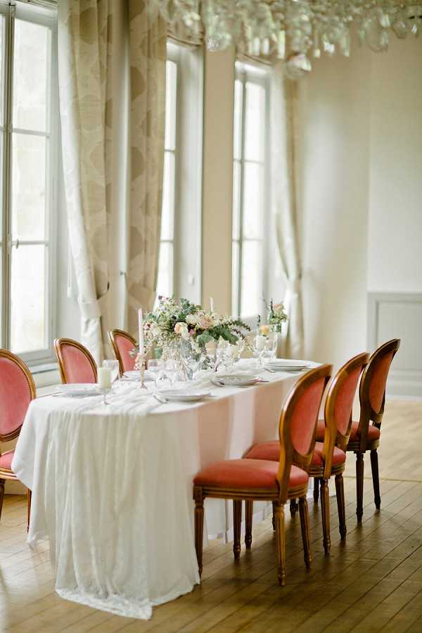 A reception tablescape detail shot taken indoors in a bright, airy room with tall French windows and patterned floor-length curtains. The rectangular dining table is dressed with a white linen tablecloth and features a low centerpiece of blush roses, ivory blooms, and soft greenery flanked by tall taper candles in glass holders. Place settings include white plates, crystal glassware, and folded napkins. The chairs are Louis XVI-style with walnut frames and terracotta-coral upholstered seats and backs, arranged on wide-plank hardwood floors. A crystal chandelier is partially visible at the top of the frame, and the overall decor palette combines white, blush, and terracotta in a classic French interior style.