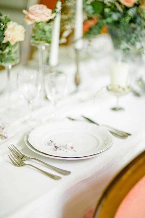 A close-up detail shot of a wedding reception table setting featuring a white linen tablecloth, stacked white china plates with a delicate purple-edged rim, and silver flatware including two forks on the left and a knife on the right. A small sprig of pale lavender and white sweet peas is placed on top of the plates as a decorative accent. Clear crystal wine glasses and a white pillar candle in a glass holder are visible in the background, alongside floral arrangements of peach roses and greenery and white taper candles in silver candlesticks. The overall decor palette is soft and romantic, combining white, peach, and pale lavender in a classic French-inspired style.