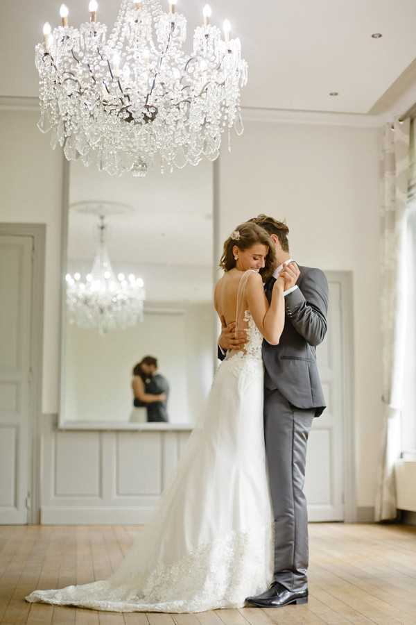 A bride and groom share an intimate embrace in a bright, white-panelled interior room with light hardwood flooring. The bride wears a fitted ivory lace gown with a low open back, floral appliqué detailing, and a trailing skirt, with her dark hair pinned up with a small hair accessory. The groom wears a mid-grey suit with a white dress shirt. A large floor-to-ceiling mirror behind them reflects their embrace from the opposite angle, creating a layered visual effect. A crystal chandelier hangs prominently in the foreground above the couple, and a second chandelier is visible in the mirror reflection. The composition is a full-length portrait shot taken indoors, likely in a chateau or manor house reception room.