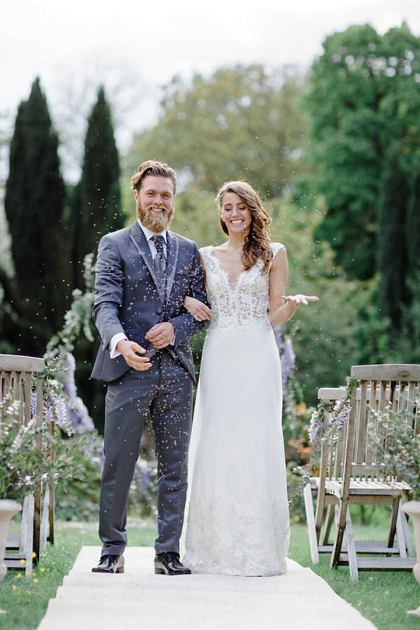 The bride and groom walk back down the aisle immediately after their outdoor garden ceremony, laughing as confetti is thrown around them. The groom wears a charcoal grey three-piece suit with a patterned tie, while the bride wears a white lace-bodice gown with a deep V-neckline and a flowing skirt. The ceremony aisle is lined with wooden folding chairs decorated with lavender-toned floral arrangements, likely wisteria or similar blooms, giving the setting a soft purple accent. The shot is a full-length portrait taken from the aisle, with a shallow depth of field blurring the garden backdrop.