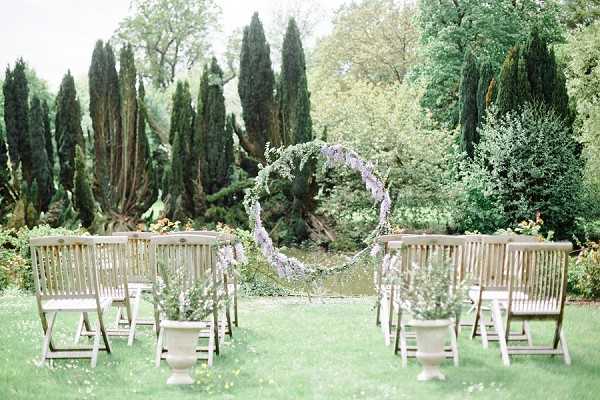An outdoor ceremony setup photographed in a garden setting, with no people present. The focal point is a large circular hoop arch decorated with trailing greenery and soft lavender-purple florals, positioned as the altar backdrop. Rows of natural wood folding chairs are arranged on either side of a central aisle on a lawn, with stone urns holding small white flowering branches placed at the aisle ends. The styling is romantic and garden-inspired, featuring a muted, soft color palette of greens, whites, and lavender. Wide shot composition capturing the full ceremony space.