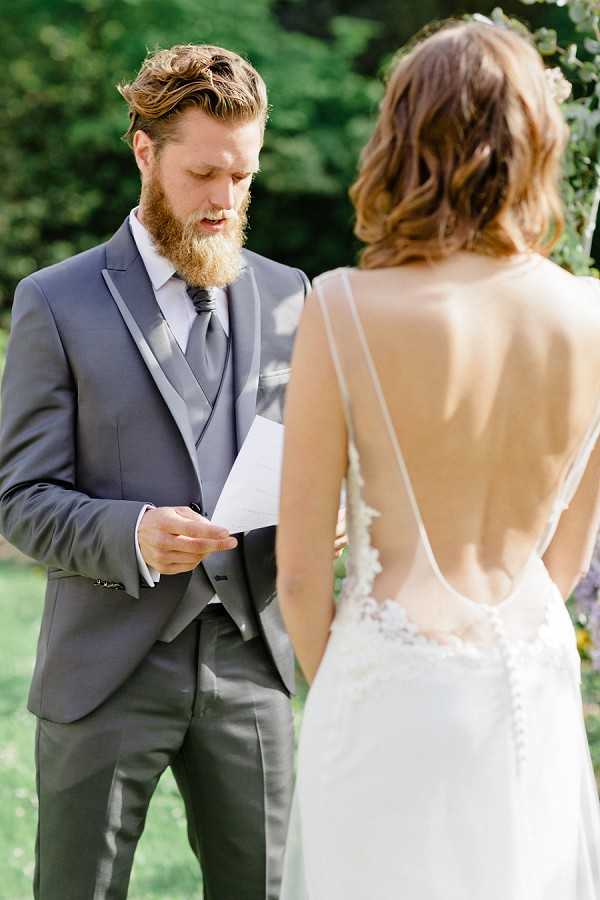 An outdoor ceremony vow exchange captured in a close-up portrait shot, showing the groom reading from a handwritten note while facing the bride. The groom has a red beard and wears a charcoal grey three-piece suit with a matching grey tie and waistcoat. The bride is seen from behind, wearing a low-open-back white gown with delicate lace appliqué detail along the back and thin spaghetti straps, her wavy brown hair worn down. A floral arch with greenery is partially visible in the background, and the setting appears to be a garden or outdoor grounds.