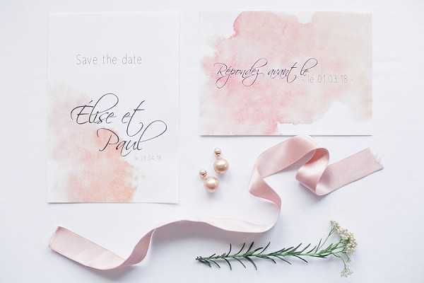 A flat lay composition showcasing a wedding stationery suite for 'Elise et Paul,' featuring a save-the-date card and an RSVP card, both printed on white paper with soft blush pink watercolor wash accents and calligraphy script typography. Styled alongside the cards are a pair of rose gold double pearl drop earrings, a loose blush satin ribbon, a small sprig of rosemary, and a few stems of white baby's breath, all arranged on a white surface. The overall stationery design palette is blush pink and white with dark charcoal lettering, reflecting a soft romantic aesthetic.