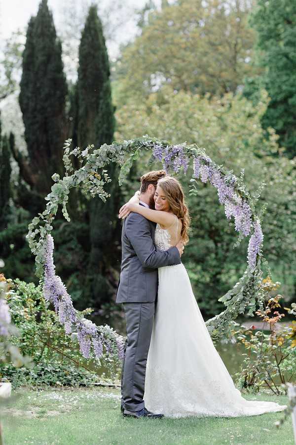 A couple portrait taken outdoors in a garden setting, with the bride and groom embracing in front of a large circular floral arch. The arch is decorated with trailing eucalyptus greenery and cascading lavender wisteria blooms. The bride wears a white A-line gown with a lace bodice and spaghetti straps, with a train extending behind her, while the groom wears a charcoal grey suit. The composition is a medium full-length portrait shot with the circular arch framing the couple centrally against a backdrop of tall cypress and mixed trees.