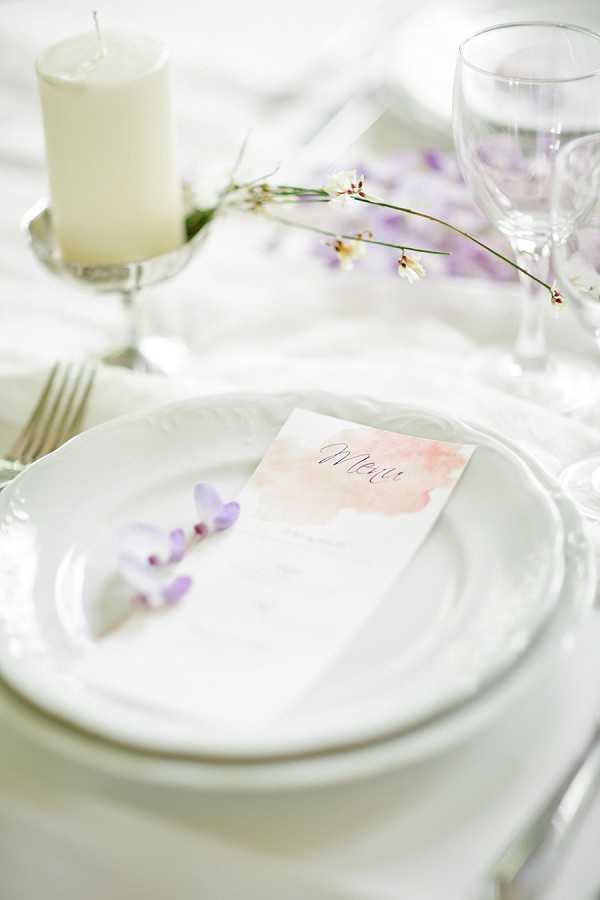 A close-up detail shot of a wedding reception place setting styled in a soft, romantic palette. The setting features stacked white ceramic plates holding a menu card with a blush pink watercolor wash design and script lettering, accompanied by a small sprig of lavender-purple orchid blooms. A white pillar candle on a silver holder, crystal glassware, and delicate white and lavender wildflower stems are visible in the background against a white linen tablecloth. The overall decor palette is white, blush, and soft lavender, consistent with a light, airy styling approach.