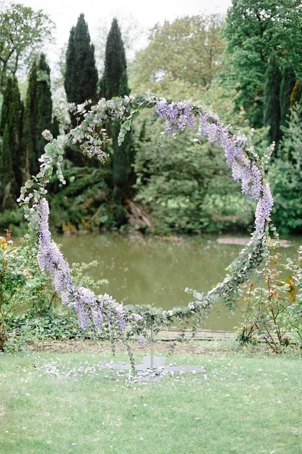 A detail shot of a circular wedding ceremony arch installed outdoors on a lawn, positioned in front of a pond. The arch is decorated with trailing lavender wisteria blooms and eucalyptus foliage, creating a full wreath-like appearance. The arch stands on a metal freestanding base, and small white flower petals are scattered on the grass below it. The overall decor palette is soft purple and green, with a romantic garden style.