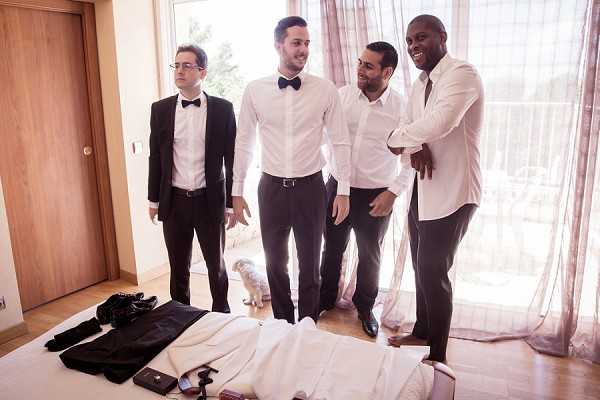 A getting-ready scene with four men standing in a bright hotel or villa bedroom, laughing and interacting casually. Two of the men are wearing white dress shirts with black bow ties and dark trousers, one is fully dressed in a black tuxedo with bow tie, and one is in a white shirt with dark trousers. A bed in the foreground holds laid-out clothing items including black trousers, a white shirt, a camera, and accessories, suggesting preparations are still in progress. A small white dog is visible near the floor in the background by large windows with sheer blush-pink curtains. The setting has a modern, clean interior with light wood flooring, and the overall styling theme is classic black-and-white formal wear. Medium-wide shot capturing the group candid moment.