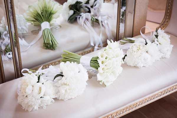 A detail/close-up shot of three white wedding bouquets arranged in a row on a cream upholstered bench or surface in front of a gold-framed mirror. The bouquets consist of white peonies, white calla lilies, and white ranunculus, each tied with white satin ribbon. The mirror's reflection shows two additional bouquets, including one with tightly gathered stems wrapped in ribbon. The overall decor palette is all-white with gold accents from the mirror frame, suggesting a classic, formal styling theme.