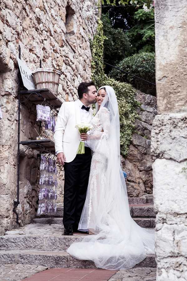 A couple portrait taken outdoors on stone steps in a narrow village alleyway, likely in a medieval hilltop village in the south of France. The groom, wearing a white dinner jacket with black trousers and a black bow tie, kisses the bride on the cheek while she smiles broadly at the camera. The bride wears a fitted long-sleeve lace gown with a cathedral-length veil and holds a compact bouquet of white calla lilies with green stems. A small retail display of lavender sachets and woven baskets is visible on a shelf mounted to the stone wall beside them, adding local character to the scene. The composition is a mid-length portrait shot, with the couple framed between rough-cut stone walls.