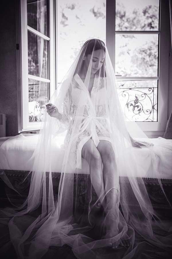 This is a black-and-white getting-ready portrait of a bride seated on the edge of a upholstered bench or bed near a large French window with ornate wrought-iron balcony details visible beyond the glass. The bride wears a short lace-trimmed robe with a satin tie belt and has her cathedral-length veil draped over her head and cascading in voluminous layers across the floor around her. She looks downward with a contemplative expression while lightly holding the edge of the veil. The image has high contrast with bright backlighting from the window creating a soft halo effect, while the foreground and corners fall into deep shadow. The composition is a full-length portrait shot from a slightly elevated angle, emphasizing the dramatic spread of the veil fabric across the floor.