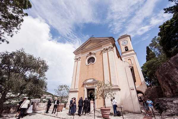 Wedding guests gather outside the entrance of a Baroque-style peach-stucco church with a clock tower, likely on the French Riviera, following or preceding a ceremony. Approximately 15–20 guests dressed in formal dark and light attire are clustered near the arched doorway, with what appears to be the couple visible among them. Two terracotta potted olive trees flank the church entrance as decorative elements. Wide-angle shot taken from a low angle looking up at the church facade, emphasizing the architecture against a partly cloudy sky. Potential venue feature image.
