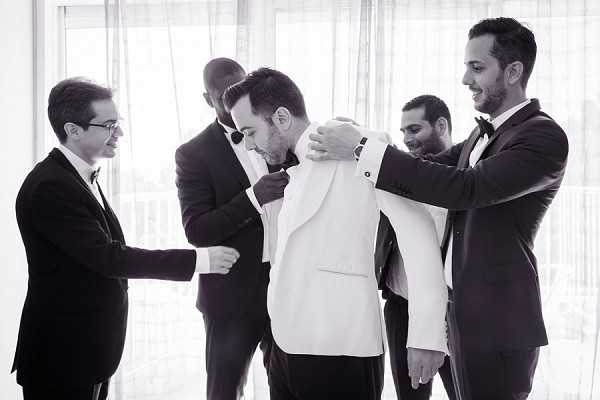 A black-and-white getting-ready scene showing a groom being helped into a white dinner jacket by four groomsmen, all dressed in dark suits or tuxedos with black bow ties. The setting appears to be an indoor room with large sheer-curtained windows providing bright, diffused backlight. The group of five men is captured in a candid, close portrait composition with the groom at the center, his head slightly bowed as the groomsmen adjust his jacket from multiple sides. The high contrast between the groom's white jacket and the dark attire of those surrounding him draws clear focus to the central figure.