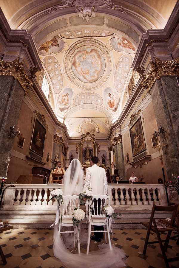 A wedding ceremony taking place inside a historic Baroque chapel with an ornate frescoed ceiling featuring classical figurative paintings and gilded architectural detailing. The bride and groom are seated with their backs to the camera at white chairs decorated with white hydrangea and eucalyptus floral arrangements tied with white ribbon, facing the altar where an officiant in gold vestments stands. The bride wears a long cathedral-length veil and a white gown, while the groom wears an all-white suit. A wide-angle shot captures the full interior from the nave to the altar, emphasizing the vaulted painted ceiling, marble balustrade, candle sconces, and large religious paintings on the side walls. The ceremony style is classic and formal, set within an ornate religious interior with warm ambient lighting. Potential venue feature image.