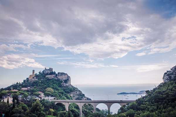 Wide-shot landscape photograph showing the hilltop village of Èze on the French Riviera, with a multi-arch stone viaduct bridge in the foreground and the Mediterranean Sea visible in the background. No people or wedding party are visible in this image. The shot appears to be a venue establishing or location shot, showcasing the dramatic clifftop architecture and surrounding scenery. Potential venue feature image.