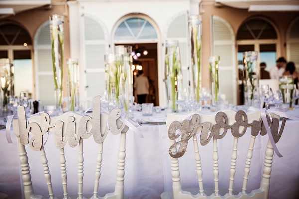 Detail shot of two white-painted wooden chairs at a wedding reception, each with a laser-cut wooden script sign reading 'bride' and 'groom' tied to the chair backs. The reception tables in the background are dressed in white linens and feature tall glass vase centerpieces holding long-stemmed white calla lilies, set against a white classical building facade with arched windows and shutters. The overall decor palette is white and pale green with a clean, classic style. A few guests are visible in soft focus in the background near the venue entrance.
