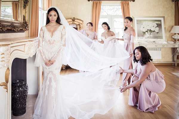 A getting-ready scene photographed indoors in an ornate room featuring a white carved fireplace, gold-framed mirror, and warm peach-toned curtains with hardwood flooring. The bride stands to the left wearing a long-sleeve, deep V-neck fitted lace gown with floral appliqué detailing and a long cathedral veil, which four bridesmaids are arranging and lifting behind her. The bridesmaids are dressed in dusty rose/mauve off-shoulder and ruffle-back satin gowns. The composition is a wide portrait-style shot capturing both the bride in the foreground and the bridesmaids working with the veil in the background, creating depth across the frame.