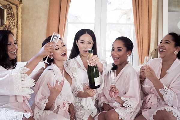 A getting-ready scene showing a bride and four bridesmaids celebrating together indoors, likely in a chateau or manor bridal suite with ornate gold-framed decor and warm drapery visible in the background. The bride, wearing a white lace-trimmed robe, is popping a champagne bottle while the bridesmaids, dressed in matching blush pink satin robes with lace trim, hold champagne flutes and react with laughter. All five women are laughing and leaning in together in a candid, lively group moment. The shot is a close-up portrait-style image with warm, natural window light.