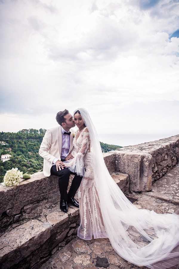 A couple portrait taken outdoors on a stone terrace or rampart with a panoramic hilltop view in the background. The groom, dressed in a white tuxedo jacket, black trousers, and black bow tie, is seated on a stone wall and kissing the bride on the cheek. The bride stands beside him wearing a long-sleeve lace and tulle fitted gown with a nude/blush underlay and a cathedral-length veil that trails along the stone floor. A small white calla lily bouquet rests on the wall to the left. The composition is a medium full-length portrait shot capturing both figures against the open sky and forested hillside backdrop.