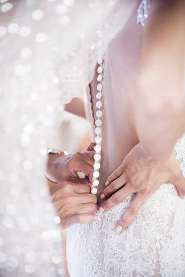 A close-up detail shot of a bride being helped into her wedding dress during the getting-ready portion of the day. Two sets of hands are visible — one fastening a row of small round buttons or button loops running along the back of an ivory lace gown. The dress features intricate lace detailing and a sheer tulle panel or veil overlapping the back closure. The background is heavily blurred with large circular bokeh highlights creating a bright, white and silver-toned backdrop. The composition is a tight macro-style detail portrait focusing on the hands and dress back.