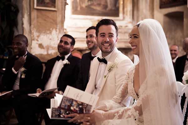 The bride and groom are seated together during an indoor religious ceremony, laughing and smiling warmly. The setting appears to be a historic chapel or church with aged plaster walls and ornate paintings visible in the background. The groom wears a white tuxedo jacket with a black bow tie and a small white floral boutonniere, while the bride wears a lace long-sleeve gown with a long veil draped over her hair. Behind them, approximately three groomsmen are visible in classic black tuxedos with bow ties and white boutonnieres, also smiling and holding ceremony booklets. The overall styling is classic and formal, and the shot is a medium close-up portrait taken from a slight angle.