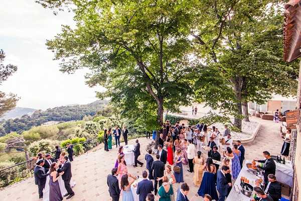A cocktail hour is taking place on a large outdoor stone terrace with panoramic views of a forested hillside valley. Approximately 50–60 guests are mingling in small groups across the terrace, with a drinks and catering station visible in the lower right corner stocked with bottles and glassware. Guests are dressed in formal attire, including navy, teal, gold, and jewel-toned dresses for women and dark suits for men. Two large mature trees provide shade over the terrace, and a curved stone balustrade borders the space. The shot is taken from an elevated wide angle, capturing the full scope of the terrace, the crowd, and the surrounding landscape. Potential venue feature image.