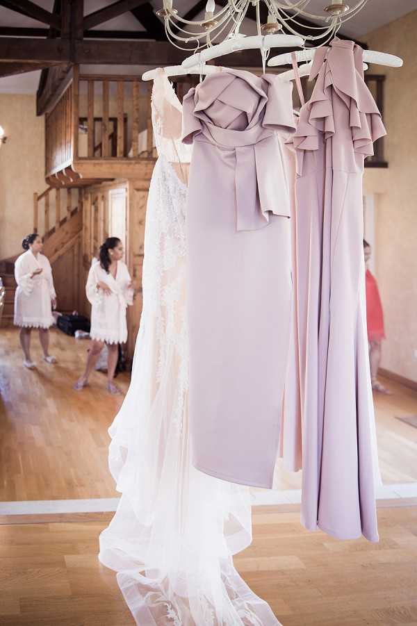 A getting-ready scene shot indoors in a room with warm wood flooring, exposed wooden beams, and a staircase, likely inside a chateau or manor house. In the foreground, a white lace wedding gown with an extended train and three mauve-lilac bridesmaid dresses — featuring sculptural rosette and ruffle neckline details — hang from a decorative chandelier on white hangers. In the soft-focus background, two women wearing white robes with lace trim stand and converse, with a third figure in a coral outfit partially visible to the right. The composition is a medium shot focused on the hanging dresses, with the bridal party preparation visible in the background.