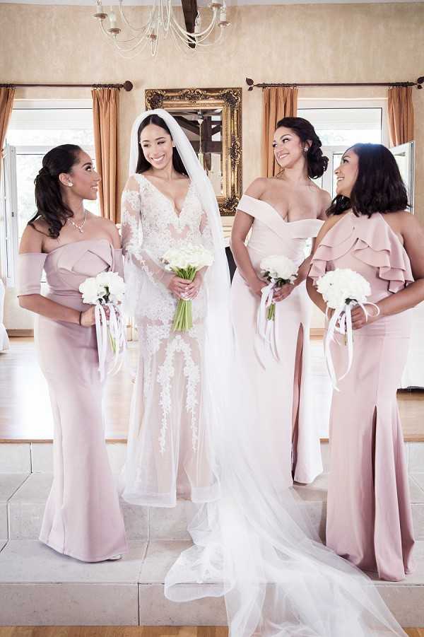 A bridal party portrait taken indoors, likely in a getting-ready room or salon of a French venue, featuring the bride and three bridesmaids. The bride wears a long-sleeve, deep V-neck lace gown with sheer illusion skirt panels and appliqué detailing, paired with a long cathedral veil, and holds a bouquet of white calla lilies with trailing white ribbons. The three bridesmaids wear floor-length blush-pink satin gowns in varied styles — one off-the-shoulder, one off-the-shoulder with structured fold detail, and one halter-neck with ruffle trim — each holding matching white calla lily bouquets with white ribbons. The room features warm cream walls, a gold ornate mirror, terracotta-toned curtains, and a chandelier overhead. The shot is a medium full-length portrait with all four women facing each other and smiling, styled in a classic, polished aesthetic with a blush and white color palette.