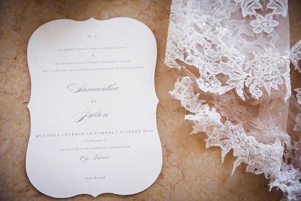 A flat lay detail shot showing a French wedding invitation card alongside a portion of a bridal veil or lace fabric. The invitation features an ornate die-cut shape with curved edges, printed in a classic serif and script typeface combination on cream or white card stock, bearing the names Samantha and Julien with a ceremony date of Saturday, July 7, 2018, at Église Notre-Dame de l'Assomption in France. To the right, a heavily embroidered Chantilly-style lace with floral and leaf motifs in ivory is partially visible, likely a veil or dress detail. Both items are laid on a warm beige marble or stone surface. The overall styling palette is ivory and cream with a classic, formal aesthetic.