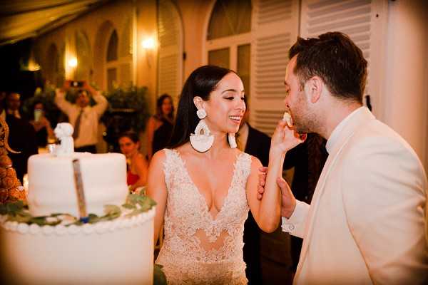 The couple is sharing a cake tasting moment during an outdoor evening reception, with the bride feeding the groom a piece of wedding cake. The bride wears a deep V-neck lace and sheer fitted gown with large white sculptural drop earrings, and the groom is dressed in a cream or off-white dinner jacket. A two-tiered white wedding cake with scalloped detailing and small green leaf accents is visible to the left. The setting appears to be a terrace or courtyard in front of a building with warm amber lighting and shuttered windows. Several guests are visible in the background, including one photographing the moment. The image is a close-up portrait shot with warm, candlelit evening tones.