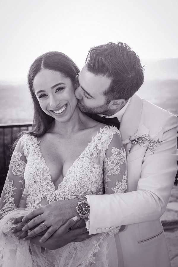 A black-and-white couple portrait showing the groom kissing the bride on the cheek while she smiles broadly at the camera. The bride wears a long-sleeve lace gown with a deep V-neckline and floral lace appliqué along the sheer sleeves, and her hair is worn down. The groom is dressed in a light-colored suit jacket with a dark bow tie, a small floral boutonnière, and a dark-dial sport watch on his wrist. The image is a close-up portrait with a soft, bright background suggesting an outdoor elevated setting such as a terrace or rooftop. The high-contrast black-and-white tones give the image a sharp, editorial quality.