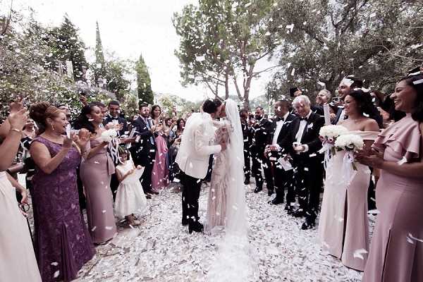 The couple shares a kiss during a confetti exit on an outdoor tree-lined path, surrounded by approximately 30 guests on either side forming a receiving line. The groom wears a white tuxedo jacket with black trousers, while the bride wears a fitted white lace gown with a long cathedral veil. White petal confetti fills the air as guests toss handfuls. Female guests wear formal attire in mauve, dusty purple, and blush pink tones, including a bridesmaid in a blush pink off-shoulder gown holding a white bouquet. The wide shot captures the full procession aisle with the couple centered in the composition.