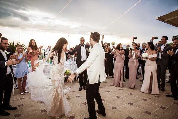 The bride and groom share a first dance on an outdoor stone terrace, surrounded by approximately 30 guests who are clapping and cheering. The bride wears a fitted, heavily embellished ivory lace gown with a full ruffled skirt and holds a small white bouquet, while the groom wears a white dinner jacket with black trousers. Bridesmaids are dressed in blush pink floor-length gowns, and several guests are photographing the moment on their phones. The reception appears to be set on a elevated terrace with white dining tables visible in the background, suggesting a venue with sea or coastal views, captured in a wide documentary-style shot at dusk.