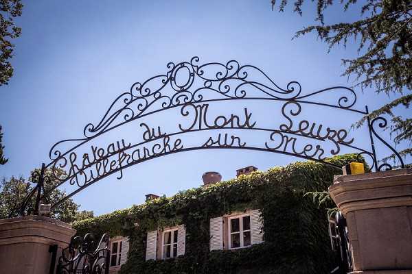 Wide-angle exterior shot looking up at the ornate wrought-iron entrance gate of Château du Mont Seure, Villefranche-sur-Mer, with the venue name displayed in decorative scrolled ironwork. The gate arches over the entrance with ivy-covered stone facade of the château building visible below, featuring white-shuttered windows. The image is framed from a low angle against a clear blue sky with tree foliage visible on either side. Potential venue feature image.