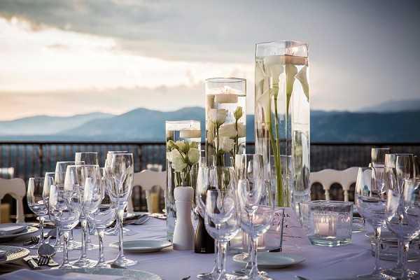 Close-up detail shot of an outdoor reception table setting on a terrace or balcony with a panoramic mountain backdrop at dusk. The centerpiece features three tall cylindrical glass vases of varying heights filled with water, white roses, white calla lilies, and submerged floating candles. The table is dressed in a white linen cloth and set with crystal wine glasses, white charger plates, and silver cutlery, with small votive candles and a place card visible. The overall decor palette is strictly white and clear glass, giving the tablescape a clean, minimalist look consistent with a modern or classic wedding style.