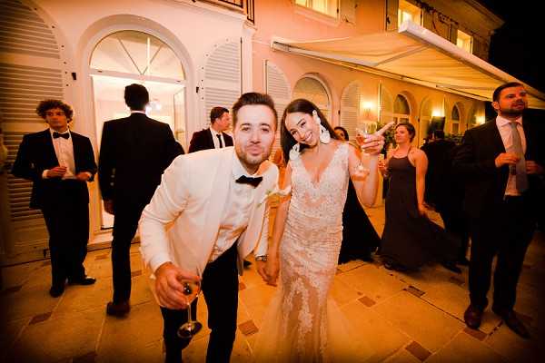 The couple poses playfully for the camera during an outdoor evening reception, both holding champagne glasses and mugging for the shot. The groom wears a white tuxedo jacket with a black bow tie, while the bride wears a fitted lace gown with a deep V-neckline and a white floral hair accessory. The setting is a stone-paved terrace outside a white classical villa or manor house with arched windows and warm interior lighting visible through the doors. Approximately six to eight guests are visible in the background, the men dressed in black tuxedos and the women in dark formal gowns, socializing under warm amber lighting that gives the image a strong orange-toned cast. Wide-angle portrait shot taken at night.