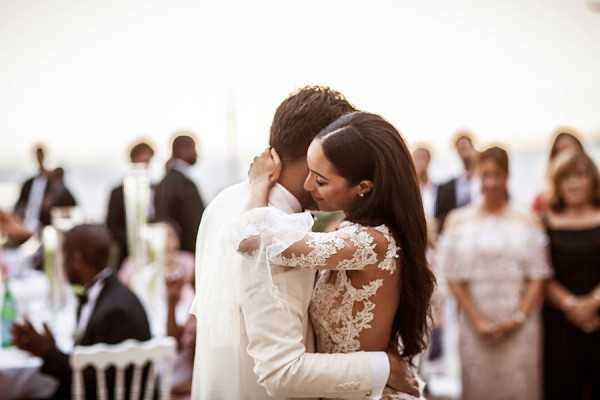 The couple shares their first dance at an outdoor reception, embracing closely with their foreheads nearly touching in an intimate moment. The bride wears a long-sleeve lace gown with floral appliqué detailing on sheer fabric and has long dark hair worn down, while the groom is dressed in a white suit. The reception appears to be set on a waterfront terrace with white Chiavari chairs and tall white floral centerpieces visible in the background, surrounded by approximately 20-30 guests watching the dance. The image is a mid-range portrait shot with a shallow depth of field, keeping the couple sharp in the foreground while the seated and standing guests blur into a warm, softly lit background.
