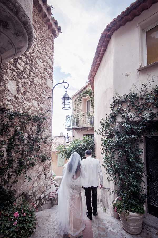 A bride and groom walk hand-in-hand away from the camera through a narrow cobblestone alley in what appears to be a historic Provençal or South of France village. The bride wears a white lace fitted gown with a long cathedral-length veil, and the groom is dressed in a white jacket with dark trousers. The alley is flanked by aged stone and rendered walls draped with climbing green vines, with a wrought-iron lantern hanging overhead and terracotta-roofed buildings visible in the background. The shot is a medium-wide portrait taken from behind, emphasizing the village architecture and the couple's path forward through the setting.
