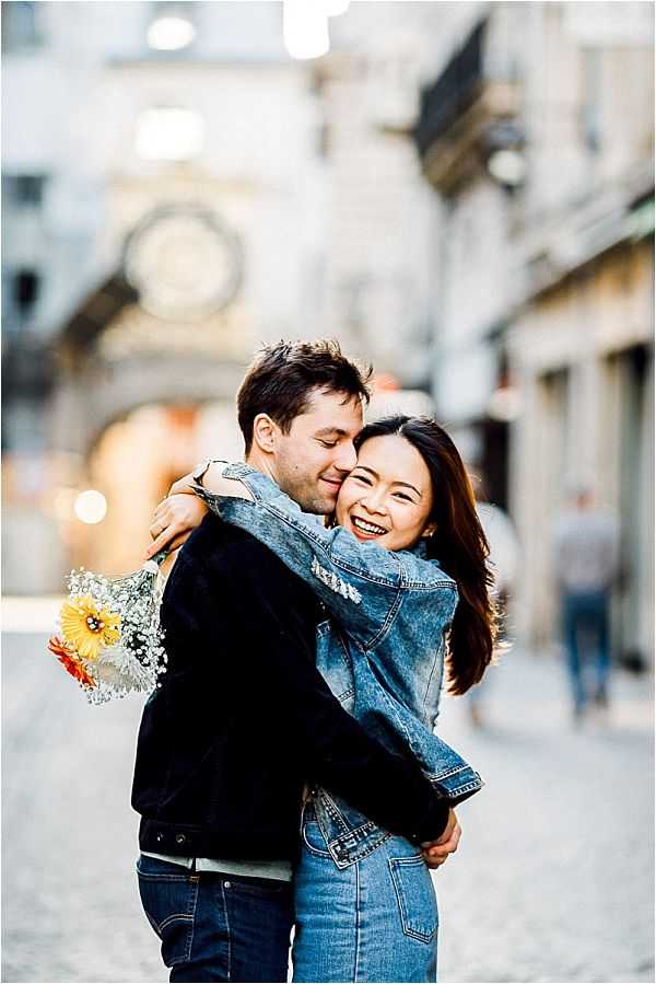 A couple portrait session set on a cobblestone street in what appears to be a French city, with blurred historic building facades and a clock visible in the background. The man, wearing a black jacket and jeans, embraces the woman from behind while nuzzling her cheek; she is laughing and wearing a denim jacket and blue jeans. She holds a small bouquet of yellow sunflowers and white baby's breath at her side. The shot is a mid-length portrait with a shallow depth of field that keeps the couple sharp against the softly blurred urban street scene.