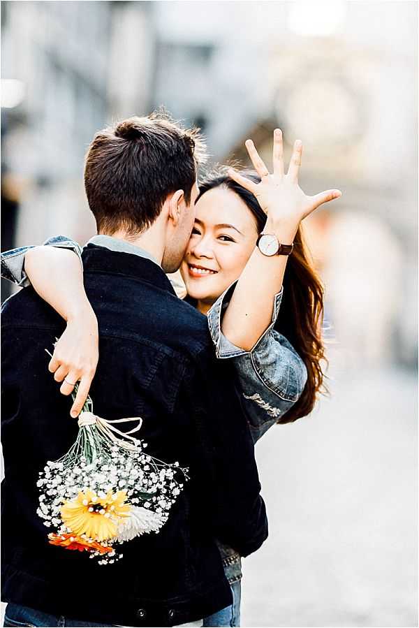 An engagement or couples portrait session shot outdoors in an urban setting with a blurred city street background. A woman smiles at the camera while embracing a man from behind, displaying her left hand prominently toward the lens to show an engagement ring; she wears a denim jacket and a round-face watch with a brown leather strap. The man, whose back is to the camera, wears a black denim jacket and holds a casual bouquet of yellow and orange gerbera daisies mixed with white baby's breath at his side. The image is a close-up portrait with shallow depth of field, captured in warm natural light.